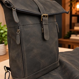 Può includere: Uno zaino grigio scuro con chiusura a rullo, finitura opaca, una tasca frontale verticale con cerniera e una cinghia centrale con fibbia in metallo color ottone. Lo zaino presenta tiretti della cerniera abbinati e spallacci neri regolabili.
