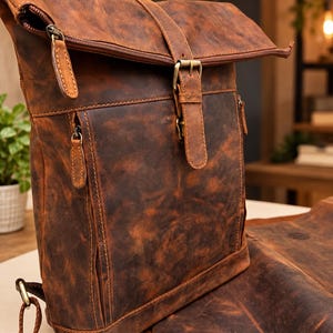 May include: A brown leather roll-top backpack featuring a distressed finish, a central vertical strap with a brass-toned metal buckle, and two front zippered pockets with matching leather pull tabs. The bag includes adjustable brown fabric shoulder straps.