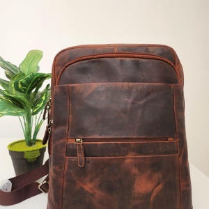 Può includere: Uno zaino in pelle marrone caratterizzato da un ampio scomparto principale con chiusura a zip, una tasca frontale più piccola con zip e un pannello frontale piatto. Lo zaino include spallacci in tessuto marrone scuro con fibbie in metallo.