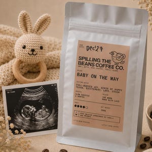 Peut inclure: Un sachet de café blanc étiqueté Spilling The Beans Coffee Co. Baby On The Way avec un poids net de 250g. L'étiquette contient des notes de dégustation, le niveau de torréfaction et l'origine. À côté se trouvent un hochet lapin, une tétine et une échographie.
