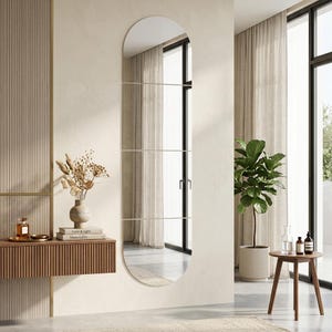 Puede incluir: Un espejo de pared grande, de forma ovalada, con la parte superior redondeada y tres barras metálicas horizontales. El espejo refleja una habitación con tonos neutros, una planta y una pequeña mesa de madera. Un elemento decorativo para el hogar.