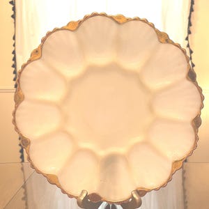Pode incluir: Um prato de servir branco com borda ondulada e borda dourada, apoiado em um suporte dourado. O prato tem uma área circular central cercada por 12 compartimentos curvos. O prato é provavelmente feito de vidro ou porcelana.