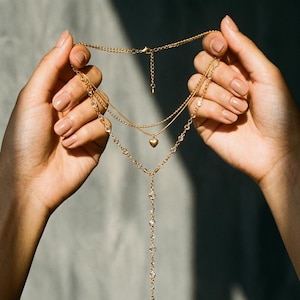 May include: A multi-strand gold necklace featuring a heart-shaped pendant and crystal accents. The necklace is displayed against a plain backdrop, highlighting its elegant design. The jewelry piece is a stylish accessory.