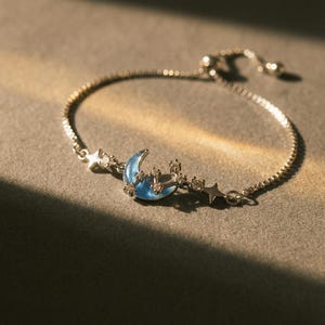 Puede incluir: Pulsera plateada con un colgante de media luna azul y detalles de estrellas. La pulsera de cadena ajustable tiene un diseño delicado, con pequeños abalorios de estrella a cada lado de la luna. La pulsera se muestra sobre una superficie gris.