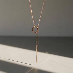 Peut inclure: Un collier délicat en or rose avec un pendentif circulaire et une barre verticale. Le collier est suspendu à une fine chaîne, créant un design minimaliste et élégant. Le bijou est présenté sur un fond neutre.