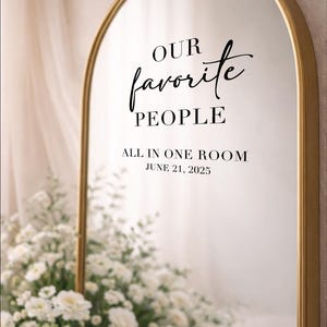 May include: A gold-framed mirror with an arched top, displaying the text "OUR favorite PEOPLE ALL IN ONE ROOM JUNE 21, 2025." The mirror is surrounded by white flowers, creating an elegant wedding or event decoration.