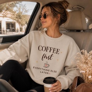 May include: Cream-colored sweatshirt with the text "COFFEE first EVERYTHING ELSE LATER" in brown script. The design includes a coffee cup graphic. The person is wearing sunglasses and holding a coffee cup.