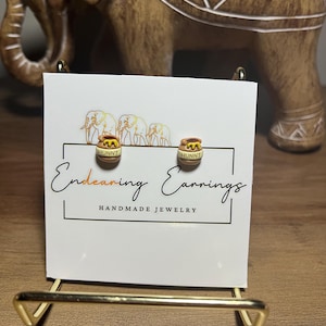 May include: A pair of handmade stud earrings featuring a honey pot design with the word "HUNNY" on the front. The earrings are displayed on a white card with the text "Endearing Earrings Handmade Jewelry". The card is held by a gold-colored metal stand.