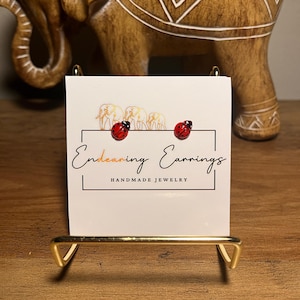 May include: A pair of ladybug earrings on a display card that reads "Endearing Earrings, Handmade Jewelry." The earrings are red with black spots. The display card is held by a gold-colored metal stand, with a wooden elephant statue in the background.