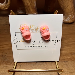 May include: Pink clog earrings with yellow and blue accents are displayed on a white card that reads "Endearing Earrings, Handmade Jewelry". The earrings are on a gold-colored stand, with a wooden elephant statue in the background.