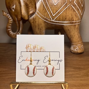 May include: Baseball-themed earrings with white and red detailing, displayed on a gold stand. The earrings feature a baseball design with red stitching. The card behind the earrings reads "Endearing Earrings MADE JEW".