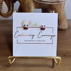 May include: A pair of cow-shaped earrings displayed on a white card with the text "Endearing Earrings Handmade Jewelry". The earrings are brown, black, and white, and are on a gold-colored stand.
