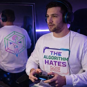 May include: A white long-sleeve shirt with the text "THE ALGORITHM HATES ME" in a purple and blue design. The shirt is worn by a person wearing headphones and holding a game controller.