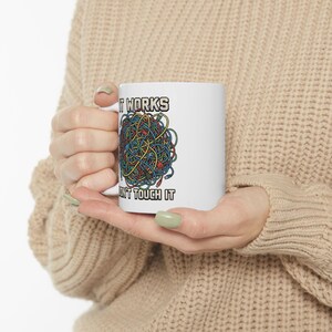 May include: White ceramic mug featuring the text "IT WORKS" above a colourful tangle of wires and the text "DON'T TOUCH IT" below. The mug is held in a person's hands.