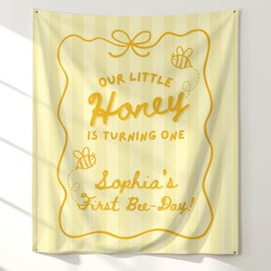 May include: A yellow banner with vertical stripes and gold lettering. The text reads "Our Little Honey is Turning One" and "Sophia's First Bee-Day!" with bee illustrations. A bow and decorative border complete the design.