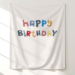 May include: White fabric wall hanging with the words "Happy Birthday" in colorful, patchwork-style letters. The letters are in red, blue, yellow, and navy blue, creating a festive and cheerful design. The fabric is slightly wrinkled.