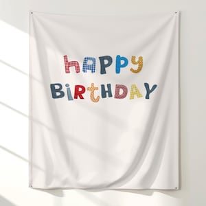 May include: White fabric wall hanging with the words "Happy Birthday" in colorful, patchwork-style letters. The letters are in red, blue, yellow, and navy blue, creating a festive and cheerful design. The fabric is slightly wrinkled.