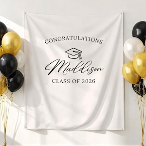 May include: A white banner with the text "Congratulations Maddison Class of 2026" is flanked by balloon bouquets in gold, black, and white. The balloons are tied with gold ribbons, creating a festive graduation scene.