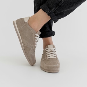 May include: A pair of taupe suede sneakers with white laces and a cream-colored heel tab. The shoes are worn with black denim jeans. The sneakers have a classic, low-profile design, suitable for casual wear.