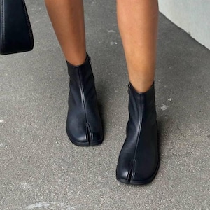 Op de afbeelding: Zwarte leren enkellaarsjes met een split-teen ontwerp. De laarzen hebben een rits aan de zijkant en een lage hak. Het schoeisel wordt van de enkels naar beneden getoond, op een grijze ondergrond.