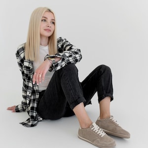 Pu&ograve; includere: Una persona indossa una maglietta bianca, jeans neri e una camicia a quadri bianchi e neri. La persona indossa scarpe da ginnastica in pelle scamosciata beige con lacci bianchi. La persona &egrave; seduta su una superficie bianca.