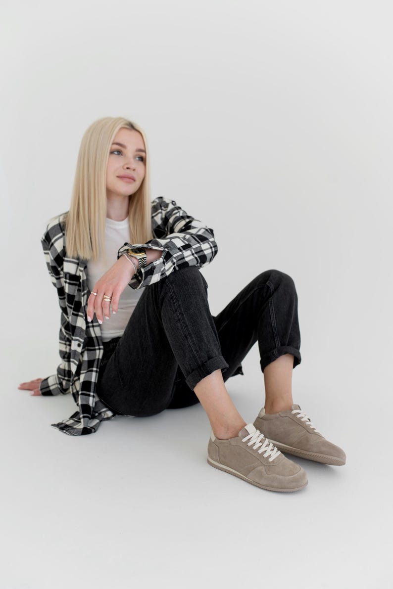 Pu&ograve; includere: Una persona indossa una maglietta bianca, jeans neri e una camicia a quadri bianchi e neri. La persona indossa scarpe da ginnastica in pelle scamosciata beige con lacci bianchi. La persona &egrave; seduta su una superficie bianca.