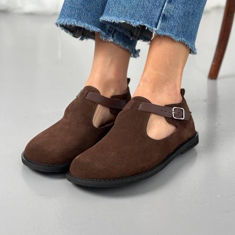May include: Brown suede ankle boots with a T-strap design and buckle closure. The shoes have a round toe and a flat sole. The boots are worn with blue denim jeans.