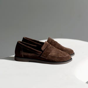 May include: A pair of dark brown suede penny loafers featuring a classic slip-on design with a rounded toe, a horizontal strap across the vamp, and a low-profile dark brown rubber sole. The shoes have a soft, matte texture and clean stitching.