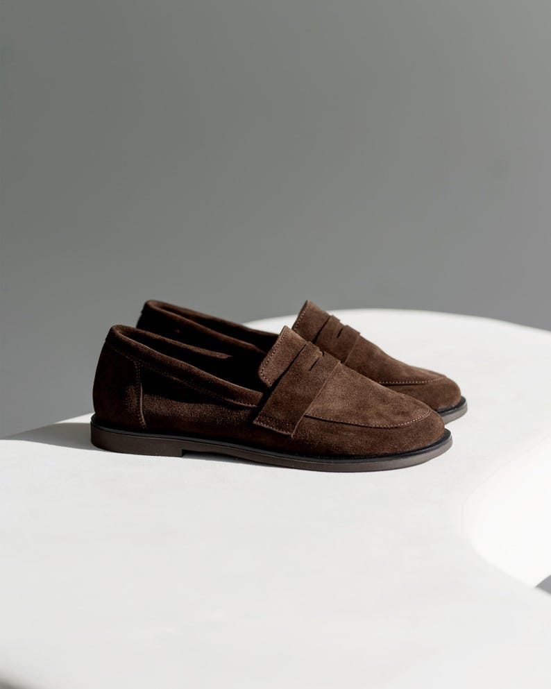 May include: A pair of dark brown suede penny loafers featuring a classic slip-on design with a rounded toe, a horizontal strap across the vamp, and a low-profile dark brown rubber sole. The shoes have a soft, matte texture and clean stitching.