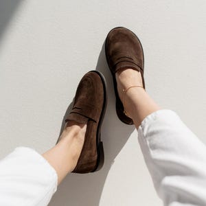 May include: A pair of dark brown suede loafers featuring a classic penny slot design across the vamp, rounded toes, and a low black heel. The shoes are worn with white trousers and a delicate gold ankle chain, showcasing a simple slip-on style.