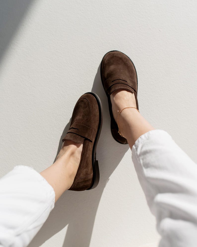 May include: A pair of dark brown suede loafers featuring a classic penny slot design across the vamp, rounded toes, and a low black heel. The shoes are worn with white trousers and a delicate gold ankle chain, showcasing a simple slip-on style.