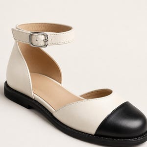 Puede incluir: Un zapato plano bicolor con cuerpo de color crema y puntera redondeada negra. El zapato incluye una correa al tobillo con una hebilla de metal plateado y una suela plana negra. El dise&ntilde;o es abierto por los lados, dejando ver el forro interior de color tostado.