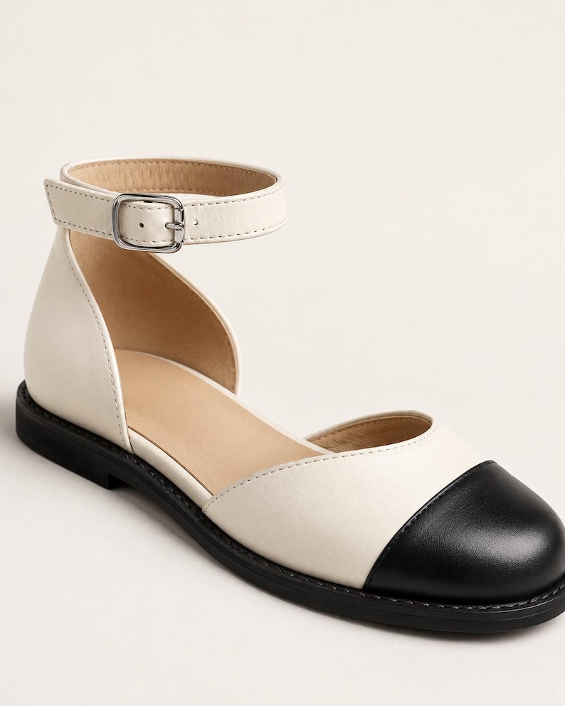 Puede incluir: Un zapato plano bicolor con cuerpo de color crema y puntera redondeada negra. El zapato incluye una correa al tobillo con una hebilla de metal plateado y una suela plana negra. El dise&ntilde;o es abierto por los lados, dejando ver el forro interior de color tostado.