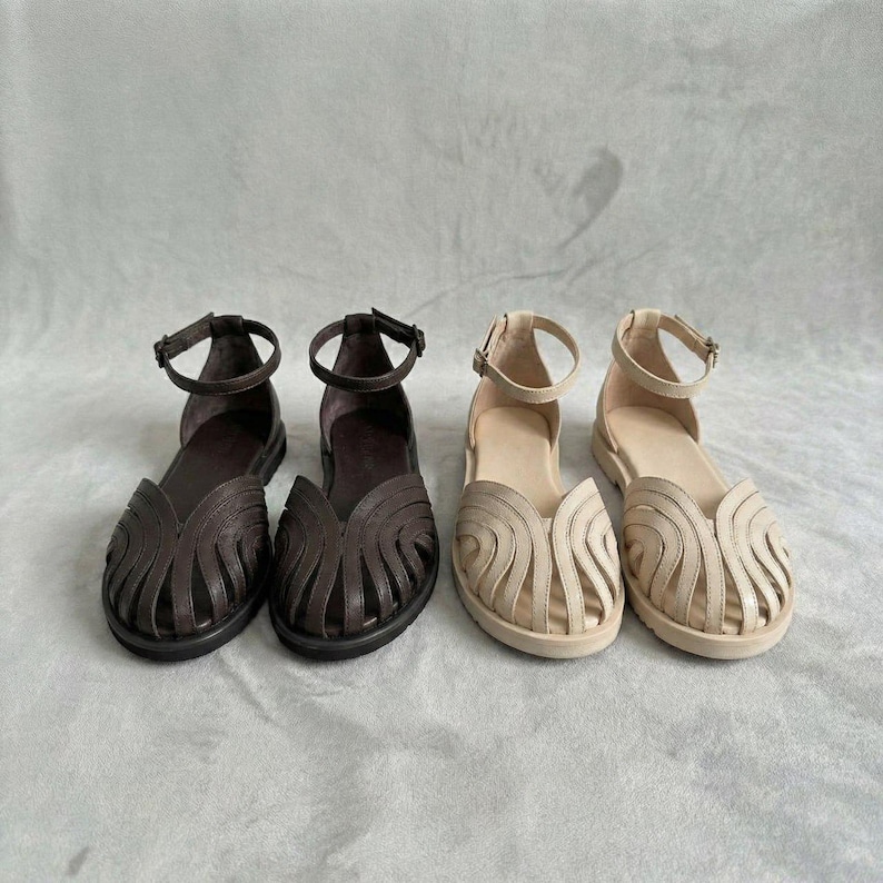 Puede incluir: Dos pares de sandalias de mujer. Un par es marr&oacute;n oscuro, el otro beige. Ambos pares tienen una correa en el tobillo y un dise&ntilde;o decorativo recortado en la parte delantera. Las sandalias son planas y parecen ser de cuero.