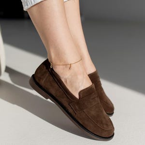 May include: A pair of dark brown suede loafers featuring a classic penny slot design across the vamp, a rounded toe, and a flat dark brown sole. The shoes are crafted from a soft, matte-textured material with visible stitching along the edges.