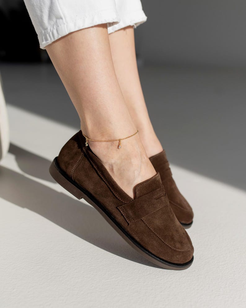 May include: A pair of dark brown suede loafers featuring a classic penny slot design across the vamp, a rounded toe, and a flat dark brown sole. The shoes are crafted from a soft, matte-textured material with visible stitching along the edges.
