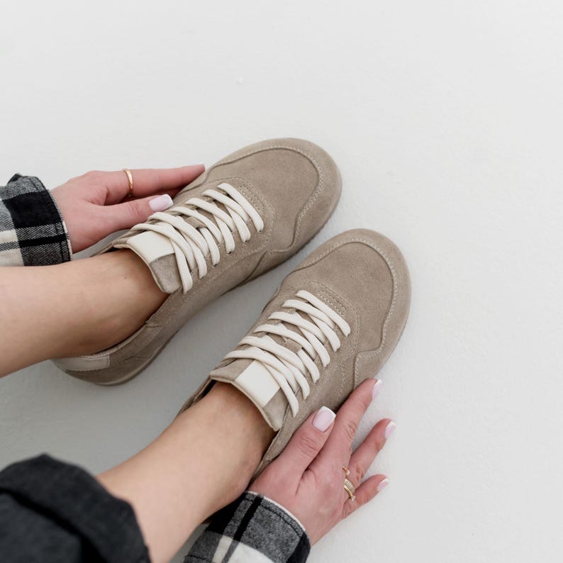 Pu&ograve; includere: Un paio di sneakers beige con lacci bianchi. Le scarpe hanno un design minimalista con una texture simile alla pelle scamosciata. I lacci sono ben legati e le scarpe sembrano nuove. Le sneakers sono mostrate su uno sfondo bianco.