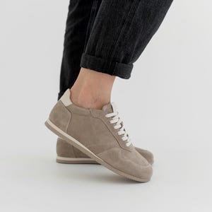 Pu&ograve; includere: Un paio di sneakers in camoscio color talpa con lacci bianchi e un rinforzo sul tallone color crema. Le scarpe a basso profilo hanno un design minimalista, adatto all'abbigliamento casual. Le suole sono color beige chiaro.
