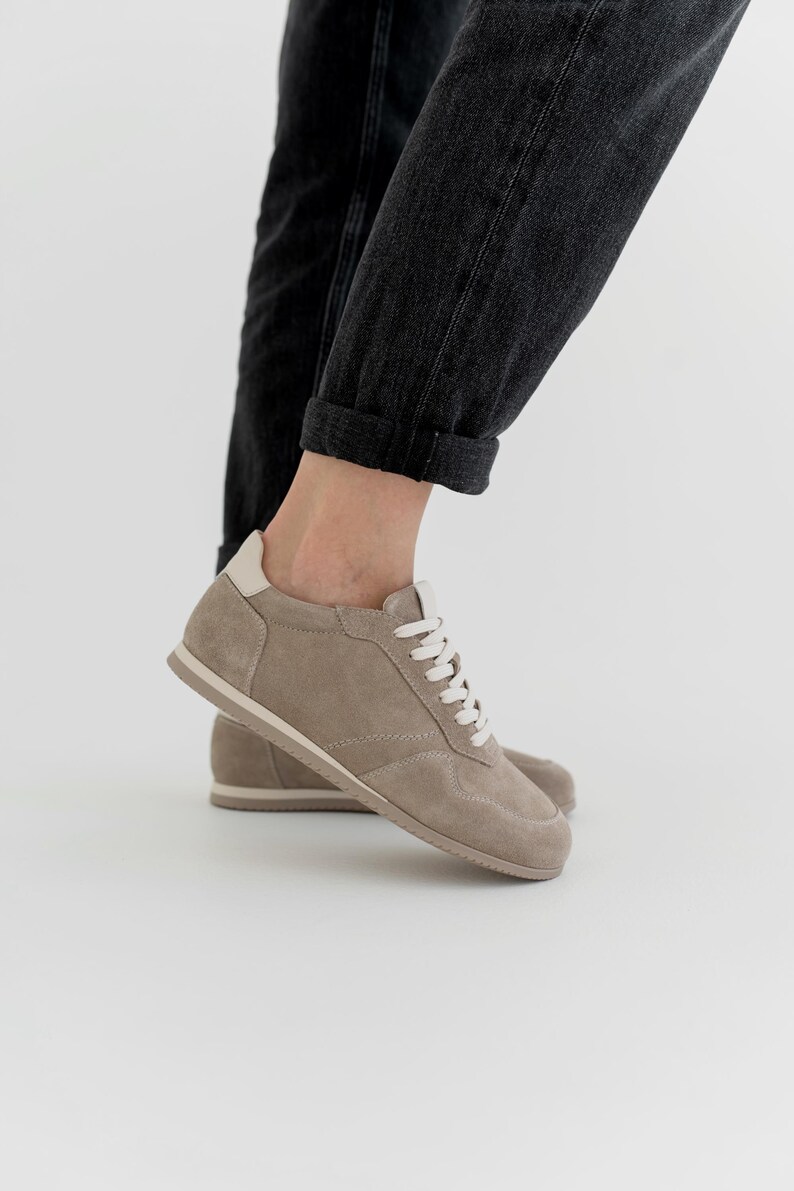 Pu&ograve; includere: Un paio di sneakers in camoscio color talpa con lacci bianchi e un rinforzo sul tallone color crema. Le scarpe a basso profilo hanno un design minimalista, adatto all'abbigliamento casual. Le suole sono color beige chiaro.