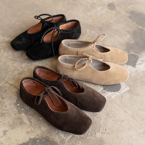 Three pairs of suede Mary Jane shoes in black, beige, and chocolate brown displayed on a concrete floor