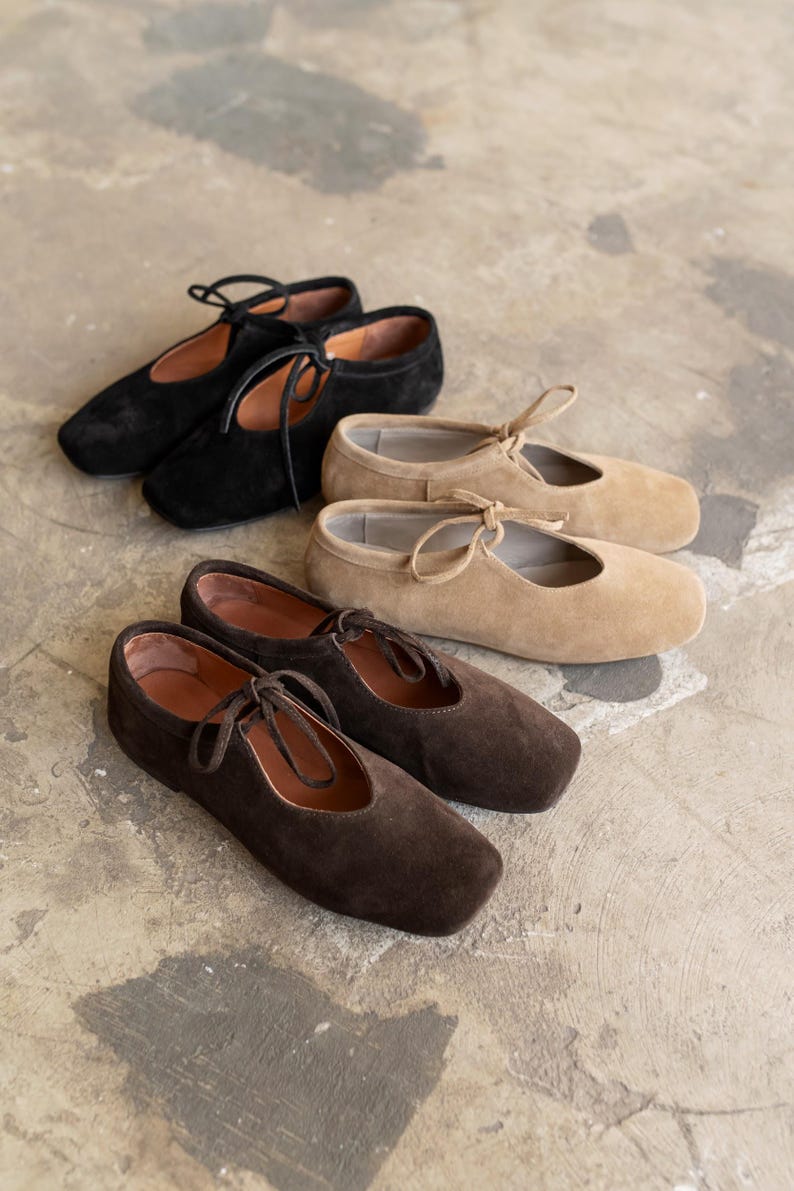 Three pairs of suede Mary Jane shoes in black, beige, and chocolate brown displayed on a concrete floor