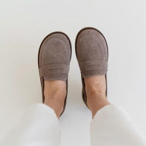 May include: A pair of gray suede penny loafers with a rounded toe and a brown sole. The shoes are shown from a top-down perspective, with the wearer's feet and white pants visible.