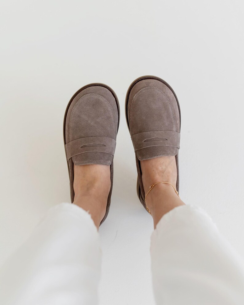 May include: A pair of gray suede penny loafers with a rounded toe and a brown sole. The shoes are shown from a top-down perspective, with the wearer's feet and white pants visible.