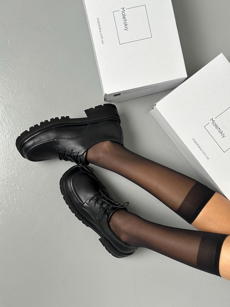 May include: Black leather lace-up boots with a chunky sole. The boots are paired with black sheer knee-high socks. Two white boxes with the brand name "Maletskiy" are in the background.