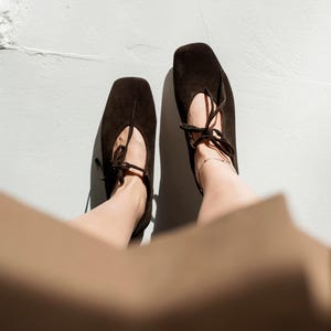 May include: Overhead shot of two dark brown suede ballet flats with square toes and delicate tie closures. The shoes are set against a white background, suggesting a focus on footwear and fashion.