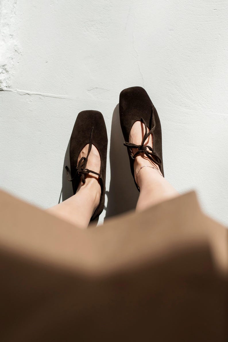 May include: Overhead shot of two dark brown suede ballet flats with square toes and delicate tie closures. The shoes are set against a white background, suggesting a focus on footwear and fashion.
