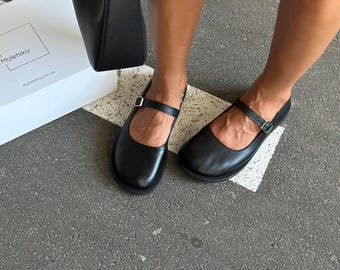 May include: Black leather Mary Jane style shoes with a buckle closure. The shoes are flat and have a rounded toe. A black handbag and a white box with the text "Maletskiy" are also visible.