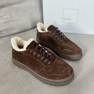 Op de afbeelding: Een paar donkerbruine suède sneakers met een crèmekleurige voering van imitatiebont, bijpassende bruine veters en een gestructureerde bruine rubberen zool. De schoenen staan voor een witte kartonnen doos met de merknaam Maletskiy erop.