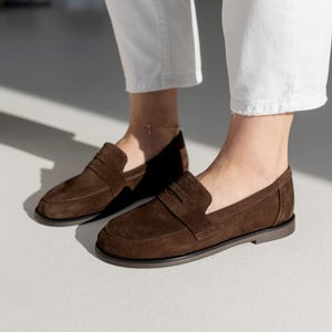 Side profile of women's chocolate brown suede loafers on a minimalist white background, showing the flat sole and classic strap