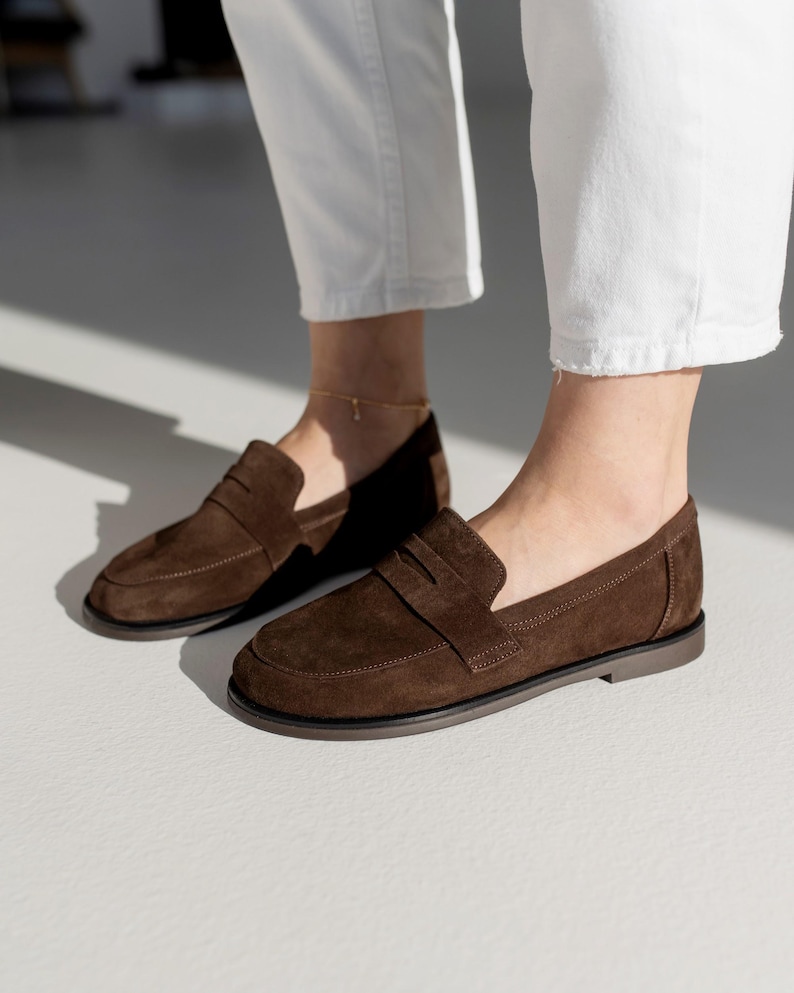 Side profile of women's chocolate brown suede loafers on a minimalist white background, showing the flat sole and classic strap